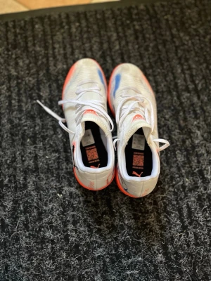 Vita och orangea Puma inomhus fotbollskor - Säljer ett par snygga vita fotbollsskor från Puma med orange sula och detaljer. Skorna har lågt skaft, klassisk snörning och är tillverkade i syntetmaterial för bra grepp och känsla på planen. Perfekta för dig som vill sticka ut på matchen!