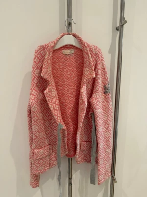 Rosa Kofta 233 - Fin rosa kofta i modell 233 från Odd Molly. Remmarna är gråa och knyts till sidan. Koftan är väl använd och har börjat bli nopprig men i övrigt är den i mycket gott skick. Säljs i befintligt skick.