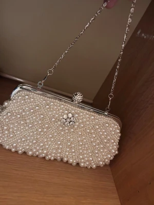Vit pärlbeströdd kuvertväska - Snygg kuvertväska täckt av vita pärlor och dekorerad med en glittrande blomdetalj framtill. Väskan har en silverfärgad metallram och en tunn kedjerem som gör den enkel att bära på axeln. Perfekt accessoar för att lyfta din outfit.