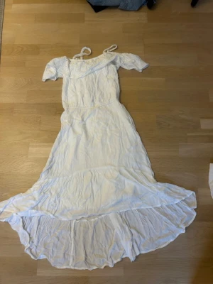 Vit lång klänning med spetsdetaljer - Söt vit långklänning med broderade spetsdetaljer upptill och axelband. Klänningen är off the shoulder och en flowy, kjol som är kortare upptill som ger en romantisk känsla. Perfekt för sommarens alla tillfällen.