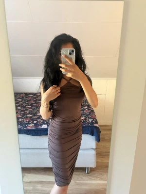 Brun midiklänning - Säljer en brun, figurnära midiklänning med oneshoulder-design och snygga rynkade detaljer längs sidorna. Klänningen har en tight passform och är tillverkad i ett stretchigt material som framhäver siluetten.