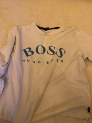 Vit sweatshirt från Hugo Boss - Vit sweatshirt med blått BOSS-tryck och texten HUGO BOSS på bröstet. Tröjan har rund halsringning och långa ärmar. Tillverkad i mjukt bomullsmaterial för en avslappnad och stilren look.