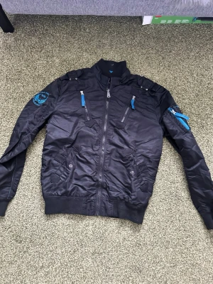 Svart bomberjacka Alpha Industries - Snygg svart bomberjacka från Alpha Industries med blå detaljer och broderad logga på ärmen. Jackan har flera dragkedjeförsedda fickor, ribbade muddar och en klassisk bomberkrage. Materialet är glansigt och ger en modern look. Helt i ny skick och strl large!