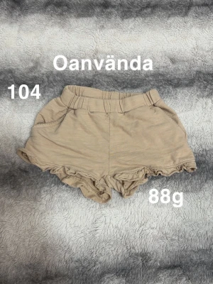 Beige shorts med volangkant - Snygga beige shorts med elastisk midja och volangkant nedtill. Perfekta för varma dagar och har en mjuk och bekväm känsla. Passar dig som gillar en söt och avslappnad stil.🌸Oanvända. Storlek 104