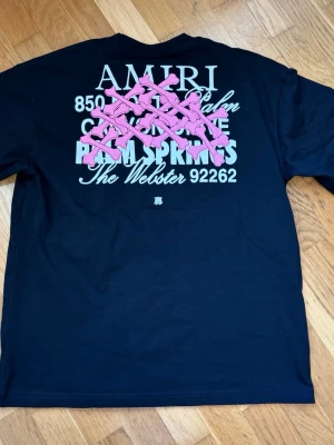 Svart AMIRI t-shirt med tryck - Svart t-shirt från AMIRI med vit och rosa text och grafiskt tryck på ryggen. Framsidan har mindre tryck med adress och logga. Klassisk passform och rund hals. Perfekt för dig som gillar streetwear med statement. Lite oversized fit