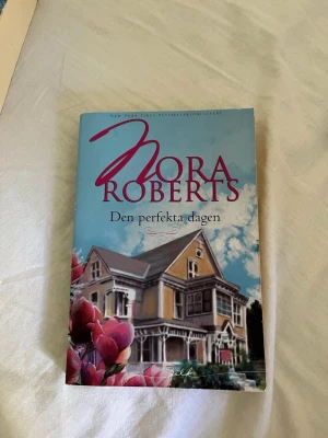 Den perfekta dagen av Nora Roberts - En romantisk roman av Nora Roberts med ett omslag i blått och rosa, prydd med en stor villa och blommor. Boken är i pocketformat och har tydliga titlar på både framsida och rygg. Perfekt för dig som gillar kärlekshistorier och lättlästa böcker.