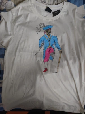 Vit t-shirt med färgglad illustration - Vit t-shirt från Diffe med en unik och färgglad illustration av en person i blå jacka, rosa byxor och hatt med fjäder. Trycket är placerat mitt på bröstet och ger tröjan en konstnärlig vibe. Perfekt för dig som vill sticka ut med din stil.