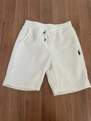 Vita shorts med snörning från Polo Ralph Lauren - Säljer ett par vita shorts från Polo Ralph Lauren med elastisk midja och snörning. Shortsen har två sidofickor och en broderad logga i svart på vänster ben. Perfekta för sommaren och riktigt sköna att bära.