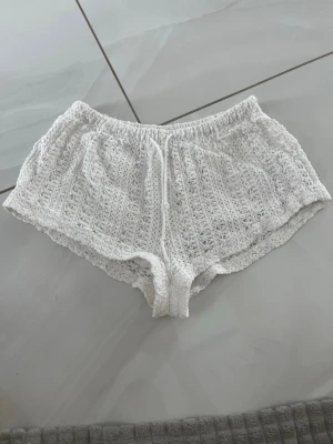 Vita virkade shorts med snörning - Säljer ett par vita virkade shorts med elastisk midja och snörning framtill. De har en luftig och avslappnad passform, perfekta för varma dagar. Materialet är mjukt och ger en bohemisk känsla.