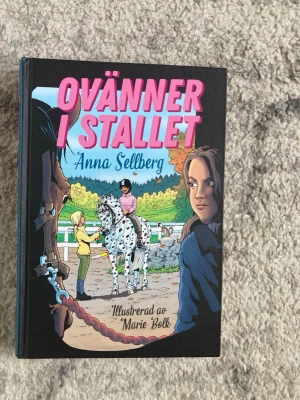 Ovänner i stallet av Anna Sellberg - En färgstark ungdomsroman med hästtema, skriven av Anna Sellberg och illustrerad av Marie Bolk. Omslaget visar en tecknad stallmiljö med hästar och ungdomar, och boken har hårda pärmar i svart med rosa och blå text.