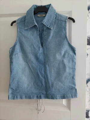 Ärmlös jeansväst från FRN - Säljer en ljusblå ärmlös jeansväst från FRN med klassisk krage och v-ringad hals. Toppen har en rak passform och dragsko i nederkant för justerbar stil. Perfekt att styla över en t-shirt eller för sig själv.