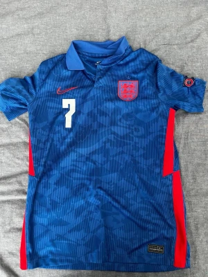 England bortatröja EM 2020 - Snygg blå England fotbollströja från Nike med nummer 7 och namnet Sancho på ryggen. Tröjan har röda detaljer på sidorna, krage och korta ärmar. England-emblem på bröstet och turneringsmärke på ärmen. Perfekt för fotbollsfans!