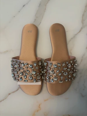 Beige sandaler med strass från H&M - Snygga beige sandaler från H&M med bred rem täckt av stora och små glittrande strass-stenar. Platt sula och öppen tå ger en trendig look. Perfekta för dig som vill sticka ut med lite extra bling i sommar.