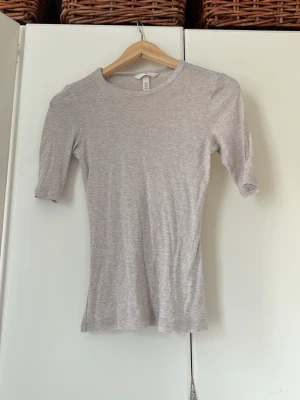 Ljusgrå ribbad topp med halvärm - En enkel och stilren ljusgrå topp med ribbad struktur och halvärm. Toppen har en rund halsringning och är figurnära i modellen, perfekt att matcha med jeans eller kjol.