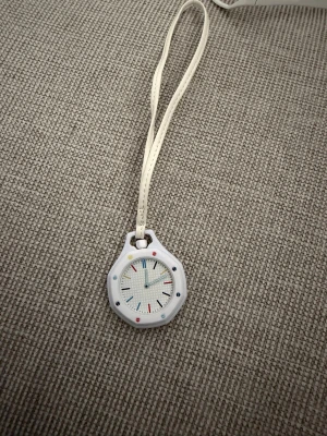 Vit klocka med färgglada detaljer - Säljer en vit klocka med silikonarmband och färgglada markeringar på urtavlan. Klockan har en modern, rund form och kan bäras både på handleden och som halsband tack vare det medföljande bandet. Perfekt accessoar för dig som gillar stilrena men lekfulla detaljer.