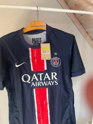 PSG matchtröja Nike herr - Säljer en officiell Paris Saint-Germain matchtröja från Nike i mörkblått med bred röd och vit rand framtill. Tröjan har PSG-logga, Qatar Airways-tryck och gul stjärna ovanför klubbmärket. Tillverkad i lätt och ventilerande material, perfekt för fotboll.