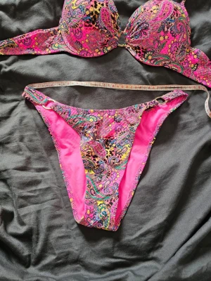 ✨ Färgglatt bikiniset med guldskimmer & high-cut (Stl S/M)✨ - Säljer ett helt oanvänt bikiniset med paisleymönster. Tyget har ett supergulligt, diskret guldskimmer som glittrar i solen. ☀️ Överdel:Vadderade kupor med bygel och knytband. Kuporna är inte så djupa, så passar bäst en A-kupa!Underdel:High-cut / high-leg-modell som går högt upp över höftbenet (ger en drömmig 80-talslook som förlänger benen!). Hög midja (25 cm).Passar bäst stl S till M (kanske mindre L pga stretchen).Underbyst: Minst 65 cm upp till 88 cm.Midja: Minst 70 cm upp till 88 cm.