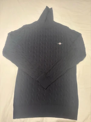 Mörkblå kabelstickad polotröja Gant - Mörkblå kabelstickad polotröja från Gant med broderad logga på bröstet. Tröjan har lång ärm, ribbade muddar och hög polokrage. Perfekt för kyliga dagar och enkel att matcha med jeans eller kjol.