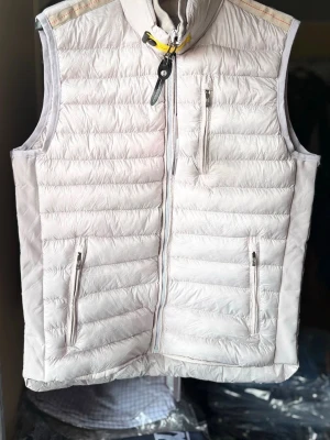 Ljusbeige dunväst från Parajumpers - Snygg ljusbeige dunväst från Parajumpers med hög krage och gul detalj vid halsen. Västen har tre dragkedjeförsedda fickor och quiltad design. Perfekt för lager-på-lager och riktigt lätt att bära. Märkespatch på insidan och orange detalj.