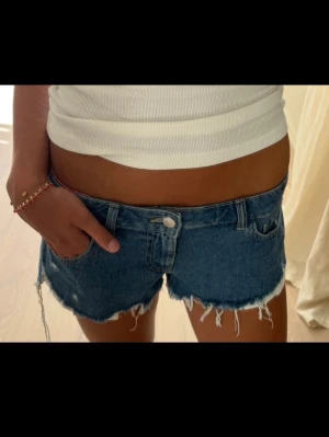 Blå denimshorts med fransig kant - Säljer ett par blå denimshorts med låg midja och rå, fransig kant. Klassisk femficksmodell med knapp och dragkedja framtill. Perfekta för varma dagar och ger en avslappnad look.