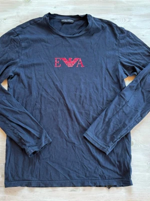 Svart långärmad topp med röd logga - Svart långärmad topp med rund halsringning och röd EA-logga framtill. Enkel och stilren design som passar till många olika outfits. Tillverkad i mjukt material för en bekväm känsla.