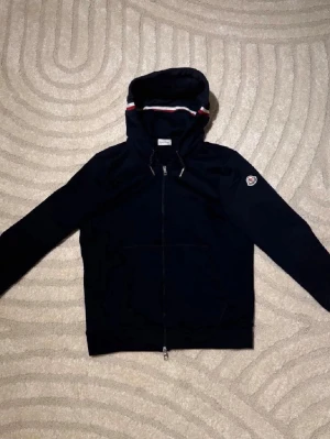 Mörkblå hoodie med dragkedja från Moncler - Snygg mörkblå hoodie från Moncler med hel dragkedja och huva. Tricolore-detalj på huvan och klassisk Moncler-logga på vänster ärm. Tillverkad i mjukt material som känns skönt mot huden. Perfekt för en avslappnad och trendig look.