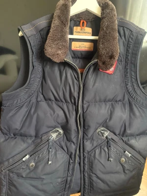 Parajumpers mörkblå dunväst - Mörkblå dunväst från Parajumpers med quiltad design, två snedställda fickor med dragkedja och knapp, samt en mjuk brun fuskpäls på kragen. Västen har dragkedja framtill och detaljer i metall. Perfekt för lager-på-lager och streetstyle.