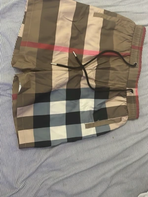 Rutiga badbyxor med dragsko - Helt nya snygga badbyxor med stort rutmönster i beige, svart, vitt, blått och rött. De har elastisk midja med dragsko och sidofickor. Perfekta för stranden eller poolen och ger en trendig look med sitt klassiska mönster.