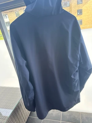 Mörkblå vindjacka med huva - Snygg mörkblå vindjacka med huva och dragkedja framtill. Jackan har två vertikala bröstfickor med dragkedja och justerbar dragsko vid huvan. Perfekt för blåsiga dagar och enkel att matcha till olika outfits.