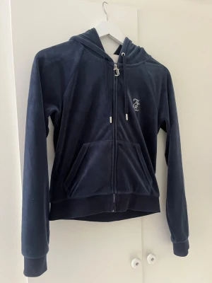 Mörkblå zip up hoodie Juicy Couture - Mörkblå mjuk hoodie från Juicy Couture. Dragkedja framtill, fickor och huva med snören. På bröstet finns en liten logga och på ryggen står det 'Juicy' i stora glittriga bokstäver. Perfekt för en chill och trendig look. 