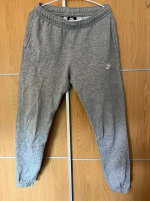 Grå mjukisbyxor från Nike - Säljer ett par klassiska grå mjukisbyxor från Nike med resår i midjan och benslut. Byxorna har sidofickor och en liten vit Nike-logga på vänster ben. Perfekta för chill eller träning, tillverkade i mjuk bomullsblandning.