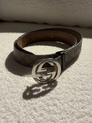Gucci beige skärp med GG-spänne - Snyggt beige skärp från Gucci med klassiskt GG-mönster och stort silverfärgat GG-spänne. Bältet har en bred rem i skinn och är perfekt för att lyfta vilken outfit som helst.