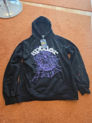 Spider hoodie - Säljer en svart hoodie med huva och ett stort lila spindelnätsmotiv på bröstet samt texten 'SPIDER' i vitt. Färgglada små detaljer finns på både fram- och ärmar. Hoodien har känguruficka och ribbade muddar. Perfekt för dig som gillar streetwear och unika prints.