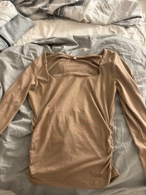 Beige långärmad topp med fyrkantig ringning - Snygg beige långärmad topp med fyrkantig ringning och rynkade detaljer på sidan. Toppen har en slim passform och är tillverkad i mjukt bomullsmaterial. Perfekt att styla med jeans eller kjol för en trendig look.