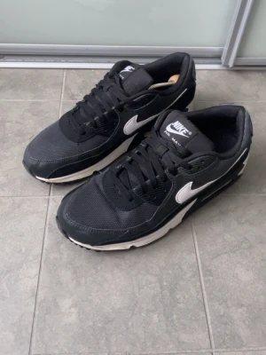 Nike Air Max 90  - Svarta Nike Air Max sneakers med vit sula och vit swoosh på sidan. Ovandelen är i mesh och syntetmaterial för en sportig look. Klassisk låg modell med snörning och vadderad plös. Perfekta för dig som gillar streetwear och stilren design.