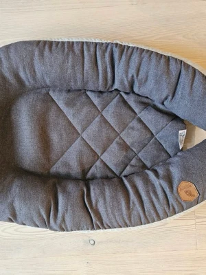 Mörkgrå babynest från Ecoviking - Säljer ett mörkgrått babynest från Ecoviking med quiltad botten och mjuka, vadderade kanter. Yttertyget är i textil och har en snygg detalj i brunt konstläder med logga. Perfekt för att skapa en mysig plats till bebisen.