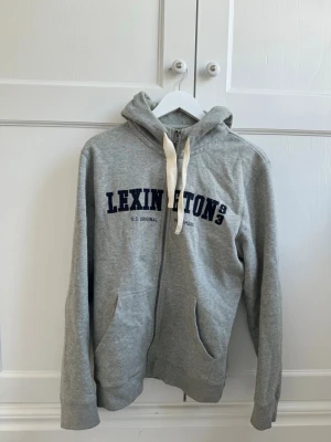 Grå zip-hoodie från Lexington - Snygg grå zip-hoodie från Lexington med mörkblå tryck på bröstet och vita dragsnören i huvan. Klassisk modell med fickor framtill och ribbade muddar. Perfekt för en avslappnad och sportig stil. Mjuk bomullsblandning för extra komfort.