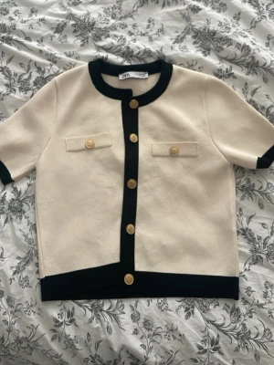 Krämvit kortärmad kofta från Zara - Stilren krämvit kofta från Zara med korta ärmar och svarta kontrastdetaljer vid krage, knappslå och nederkant. Guldknappar framtill och dekorativa fickor på bröstet ger en lyxig känsla. Perfekt till jeans eller kjol för en chic look.