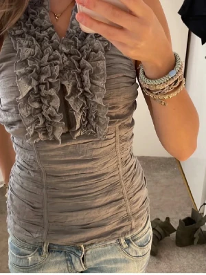 Grå volang topp med omlottkrage - Snygg grå topp med volangdetaljer längs omlottkragen. Toppen är ärmlös och har en tight passform med rynkade sömmar som ger en cool struktur. Perfekt att styla med jeans eller kjol för en trendig look.