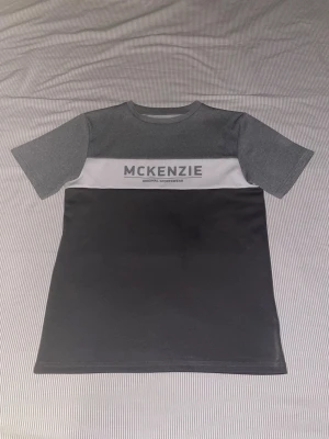 Grå McKenzie t-shirt med logga - Snygg t-shirt från McKenzie i olika grå nyanser med vit panel och stor logga över bröstet. Klassisk rak passform och korta ärmar. Tillverkad i mjuk polyester som känns skön mot huden. Perfekt för en sportig och avslappnad stil.