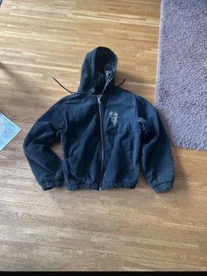 Svart hoodie från Stüssy Workgear - Svart hoodie från Stüssy Workgear med dragkedja framtill och broderad logga på bröstet. Hoodien har elastisk midja och ärmar samt två fickor fram. Tillverkad i vadderat material som ger extra värme. Perfekt för en streetwear-look.                          Säljer för att jag köpt en ny, försöker få bort denna. Pris kan diskuteras 