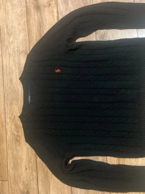 Svart kabelstickad tröja Polo Ralph Lauren - Kabelstickad svart tröja från Polo Ralph Lauren med klassisk röd logga broderad på bröstet. Tröjan har rund halsringning och långa ärmar. Perfekt för dig som gillar stilrena och tidlösa plagg.