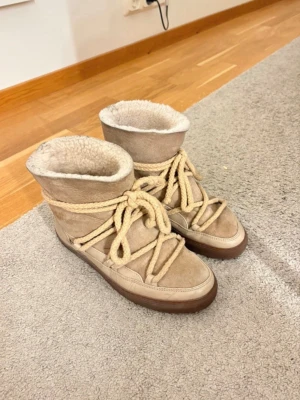 Inuikki beige - Säljer ett par beige kängor med mjukt fårskinnsfoder och grova snören runt hela skon. Yttermaterialet är mocka och läder, sulan är brun och robust. Perfekta för kalla dagar och riktigt sköna att ha på sig. Mockan är lite förstörd, därav billigt pris! Lädern lite sprucken men fortfarande varma och sköna och vattentåliga