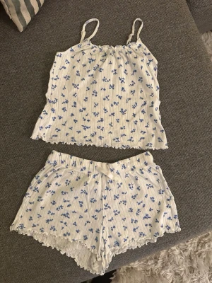 Blommigt ribbat set med linne och shorts - Sött tvådelat set med linne och shorts i vitt ribbat tyg med små blå blommor. Linnet har smala axelband och båda delarna har vågig kant nedtill. Perfekt som pyjamas. 