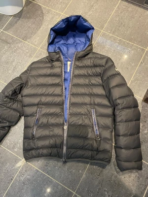 Svart dunjacka från Colmar - Snygg svart dunjacka från Colmar med blått foder och huva. Jackan har två dragkedjeförsedda fickor framtill och quiltad design. Perfekt för kalla dagar när du vill hålla stilen och värmen.