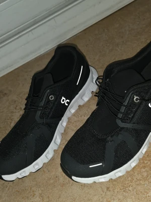 Svarta On Cloud sneakers - Snygga svarta On Cloud sneakers med vit sula och reflekterande detaljer. Ovandelen är i mesh och syntet för bra andningsförmåga. Skorna har elastiska snören och en unik, dämpad sula som ger grym komfort. Perfekta för dig som gillar sportig och modern stil.