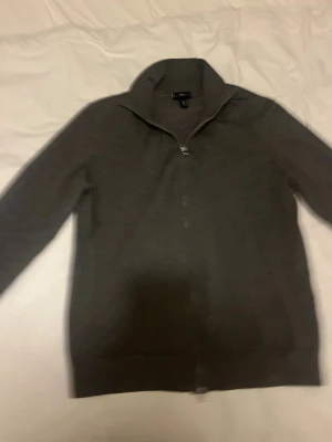 Mörkgrå ziptröja från H&M - Mörkgrå ziptröja från H&M med hög krage och hel dragkedja framtill. Tröjan har långa ärmar och ribbade muddar vid ärmslut och nederkant. Tillverkad i mjukt och tunt material, perfekt för lager på lager.