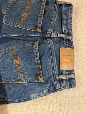 Nudie Jeans blå raka jeans W24 L30 - Säljer ett par klassiska blå jeans från Nudie Jeans med raka ben och snygga kontrastsömmar. Jeansen har hög midja, fem fickor och Nudie-logga på bakfickan. Tillverkade i slitstark bomull med orange detaljer och patch bak i midjan. W24 och L30