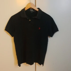 Svart pikétröja från Polo Ralph Lauren - Klassisk svart pikétröja från Polo Ralph Lauren med korta ärmar och krage. Tröjan har två knappar vid halsen och den ikoniska röda logotypen broderad på bröstet. Perfekt till jeans eller shorts för en clean look.