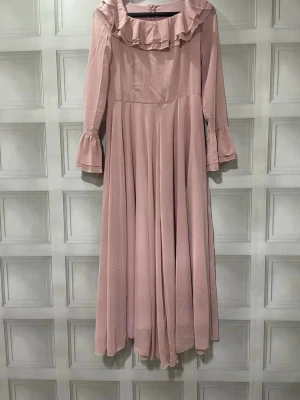 Ljusrosa långklänning med volang - Romantisk ljusrosa långklänning med volangdetaljer vid halsringningen och ärmsluten. Klänningen har långa ärmar och ett luftigt, svepande fall som ger en elegant siluett. Perfekt för dig som gillar en feminin och klassisk stil.